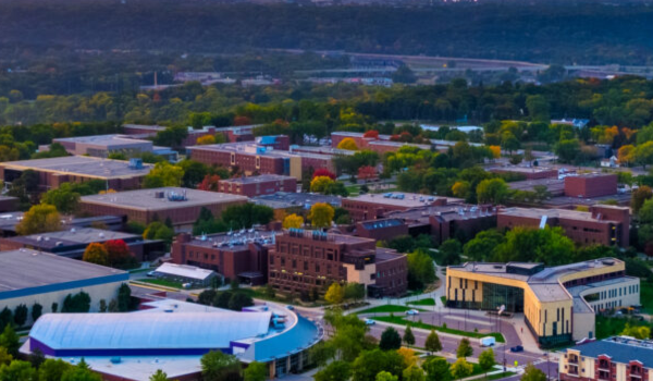 Aerial view of a university campus with multiple brick buildings, green spaces, and a distinctive modern structure in the foreground, where students and faculty thrive and proclaim: "Let's Connect!" This vibrant oasis is surrounded by trees offering inspiration for collaboration.