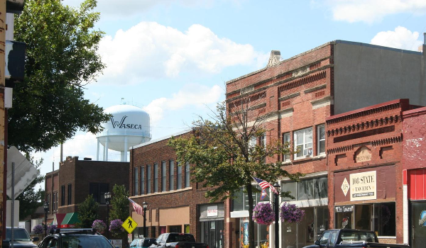 Small town street scene with brick storefronts, trees, and cars lining the road. A visible water tower labeled "Waseca" stands proudly under a partly cloudy sky, inviting locals and visitors alike to explore and say, "Let's Connect.